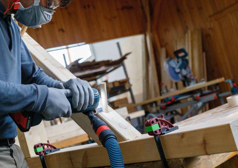 Eine Person, die Schutzausrüstung trägt, benutzt ein Elektrowerkzeug, um einen Holzbalken in einer Holzwerkstatt zu schleifen, mit verschiedenen Werkzeugen im Hintergrund.