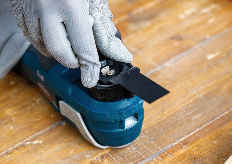 Eine behandschuhte Hand befestigt eine schwarze Klinge an einem blauen Multifunktionswerkzeug mit Oszillationsbewegung auf einer Holzoberfläche.