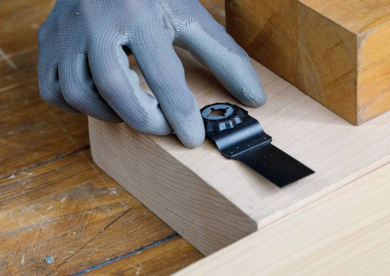 Eine behandschuhte Hand stellt ein schwarzes, gezahntes Sägeblatt zurecht, das auf einem hellholzernen Brett auf einer Holzoberfläche ruht, bereit zum Schneiden.