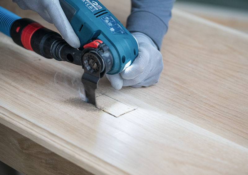 Eine Person mit grauen Handschuhen verwendet ein blau-schwarzes elektrisches Werkzeug, um eine rechteckige Form in eine Holzoberfläche zu schneiden.