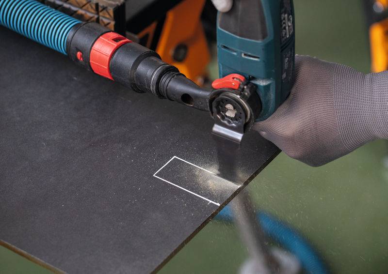 „Eine behandschuhte Hand benutzt ein Elektrowerkzeug, um eine rechteckige Form in einem schwarzen Material zu schneiden, wobei Sägespäne um den Schnittbereich sichtbar sind.