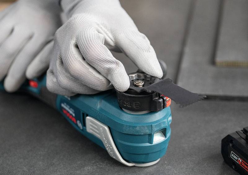 Eine Person in weißen Handschuhen befestigt ein schwarzes Schneidblatt an einem blauen Elektrowerkzeug auf einer grauen Oberfläche und bereitet es zur Verwendung vor.