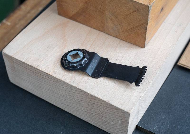 Eine Mehrzweck-Sägeblatt ruht auf einem Holzblock und zeigt seine gezahnte Kante und seinen Befestigungsmechanismus. Die Umgebung deutet auf eine Werkstatt hin.