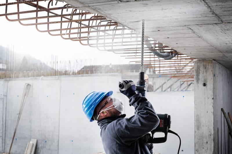 Ein Bauarbeiter in einem blauen Helm und einer Maske benutzt eine Elektrobohrmaschine an einer Betondecke, umgeben von freiliegenden Stahlbewehrungen.