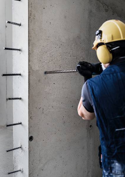 Ein Bauarbeiter in Schutzausrüstung bohrt in eine Betonwand, umgeben von vorstehenden Bewehrungsstäben, was auf Bauarbeiten hinweist.