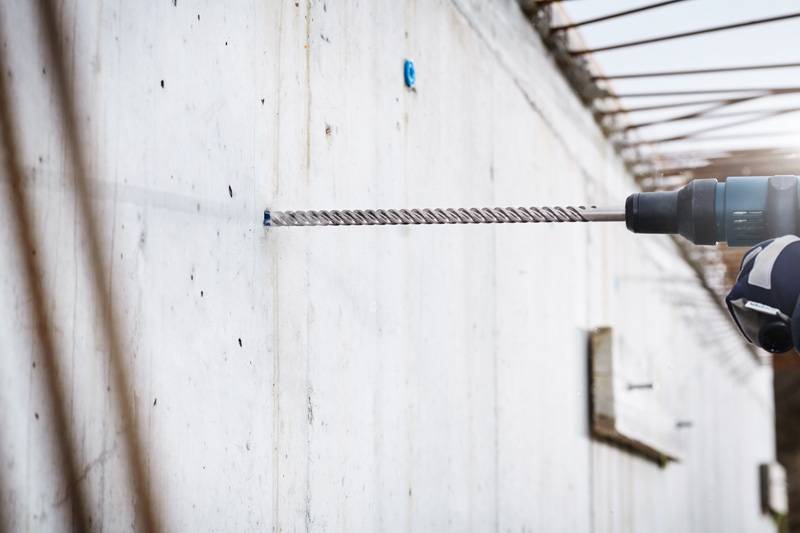 Eine Person benutzt einen großen Elektroschrauber, um ein Loch in eine Betonwand zu bohren. Metallstangen und ein kleiner blauer Marker sind an der Wand sichtbar.