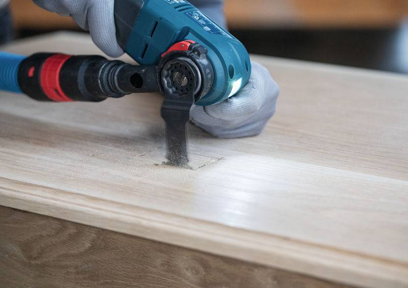 Eine Person nutzt ein blaues Elektrowerkzeug, um eine rechteckige Form in ein Holzbrett zu schneiden und trägt dabei Handschuhe zum Schutz.