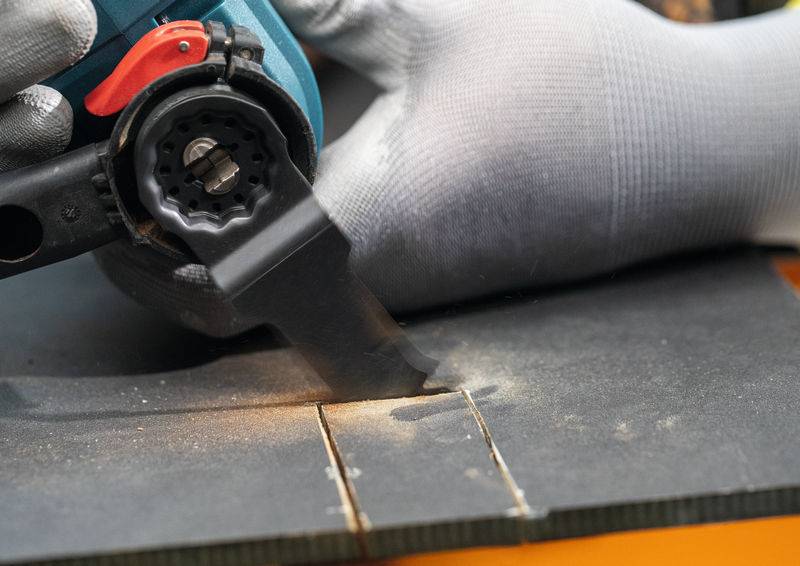 Eine Person mit Handschuhen verwendet ein elektrisches Elektrowerkzeug, um Rillen in ein schwarzes Material zu schneiden, wobei Staub und präzise Linien entstehen.