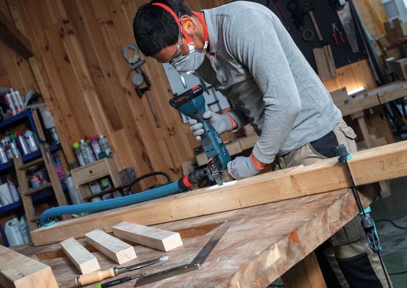 Eine Person benutzt eine Elektrobohrmaschine an einem Holzbalken in einer Werkstatt und trägt Schutzausrüstung wie Ohrenschützer und eine Maske. Holzbretter und Werkzeuge sind sichtbar.