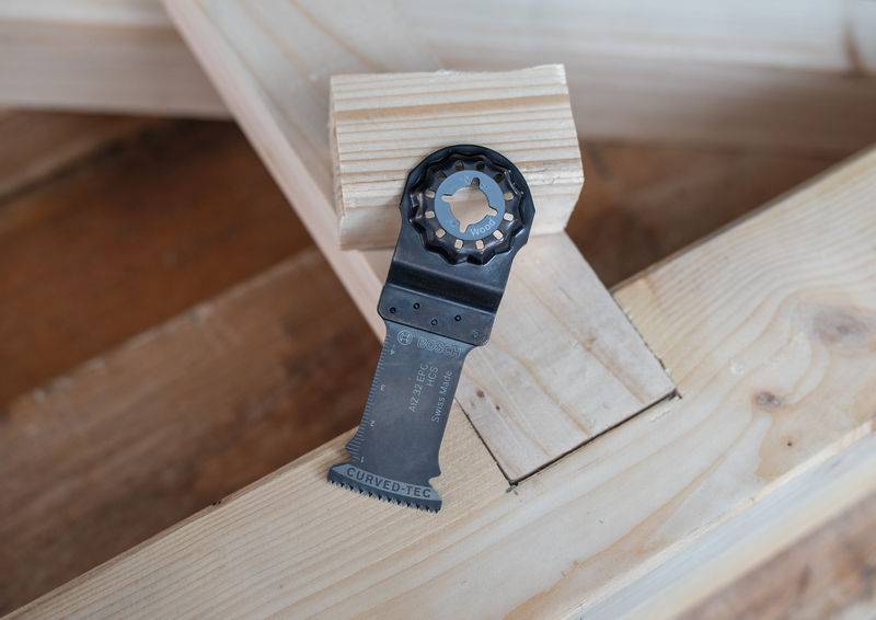 Ein Multitool-Blatt auf Holzstücken ruhend, mit sichtbaren Zähnen und Befestigungsende, in einer Werkstattumgebung eingebettet.