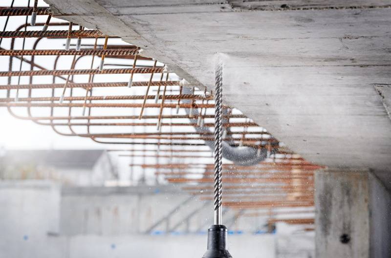 Ein Bohrer durchdringt eine Betonplatte mit freiliegender Bewehrung, was auf laufende Bau- oder Renovierungsarbeiten hindeutet.