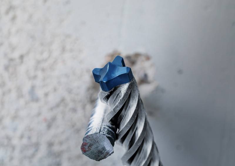Eine Nahaufnahme eines Bohrers mit einer blauem sternförmigen Spitze, der in eine Betonwand eingeführt ist, zeigt spiralförmige Nuten und Staub auf dem Bohrer.