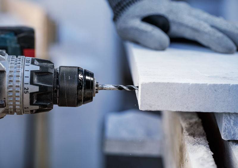 Eine Person in Handschuhen nutzt eine Elektrobohrmaschine, um ein Loch durch eine weiße Steinplatte zu bohren, wobei der Fokus auf Präzision und Kontrolle liegt.