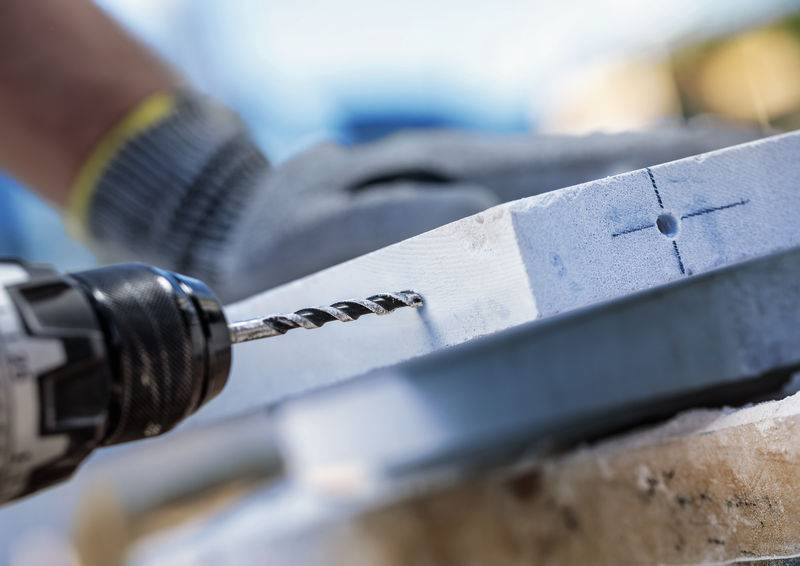 Eine Person mit Handschuhen nutzt einen Bohrer, um ein Loch in ein Holzbrett zu bohren, das mit Kreuzlinien zur Gewährleistung der Genauigkeit gekennzeichnet ist.