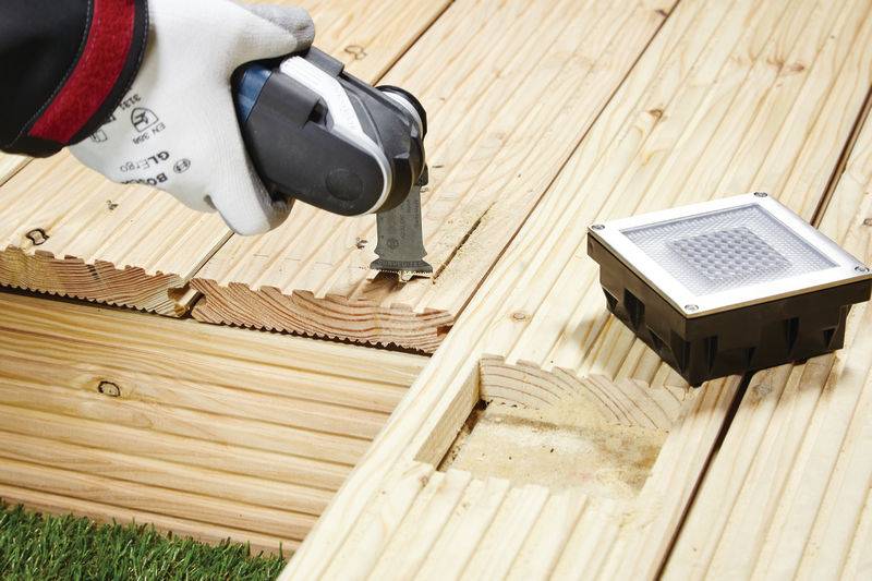 Eine Person mit Handschuhen verwendet ein oszillierendes Multifunktionswerkzeug, um ein Holzdeck zu trimmen, wobei eine Aussparung für ein quadratisches Solar-Deckenleuchte in der Nähe sichtbar ist.