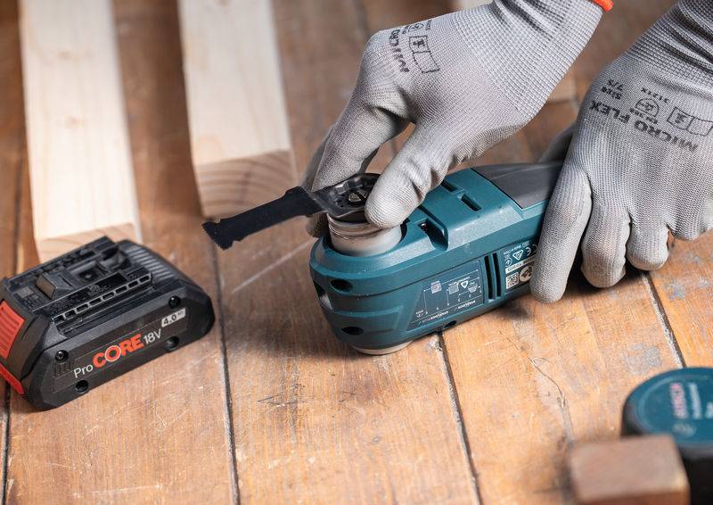 Eine Person mit Handschuhen benutzt ein Elektrowerkzeug auf einer Holzoberfläche neben einem Akku, was auf eine Bau- oder Holzbearbeitungstätigkeit hindeutet.