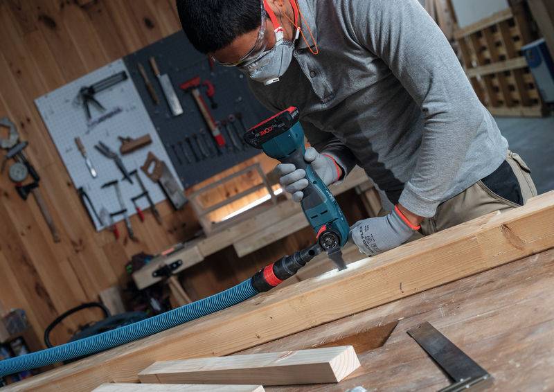 Eine Person in Schutzausrüstung bedient eine Bohrmaschine in einer Holzbearbeitungswerkstatt. Werkzeuge und Holzbretter sind im Hintergrund sichtbar.