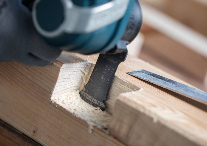 Eine Person, die ein Elektrowerkzeug verwendet, um eine rechteckige Kerbe in ein Holzbrett zu schnitzen, mit sichtbaren Holzspänen und einem Meißel in der Nähe.