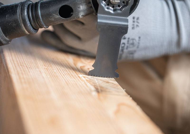 Eine Nahaufnahme einer behandschuhten Hand einer Person, die ein oszillierendes Werkzeug zum Zuschneiden eines Holzstücks verwendet, gezeigt in einer Werkstattumgebung.