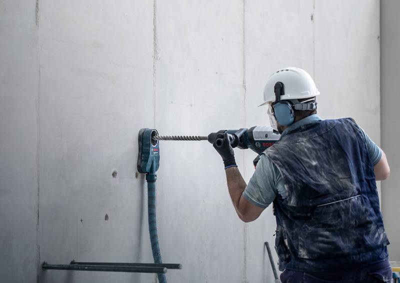 Ein Bauarbeiter in Schutzausrüstung verwendet einen Bohrer an einer Betonwand, was darauf hindeutet, dass Bau- oder Renovierungsarbeiten im Gange sind.