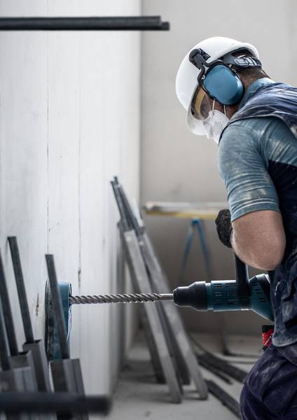 Ein Bauarbeiter in Schutzausrüstung bedient einen Bohrer an einer weißen Wand, umgeben von Metallstangen in einem teilweise fertiggestellten Raum.