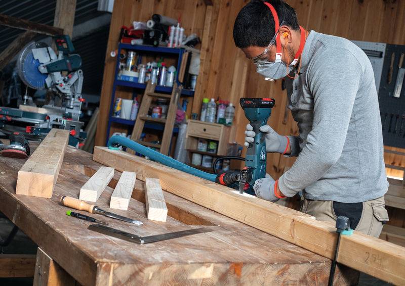 Eine Person, die eine Maske und Schutzbrille trägt, bohrt in ein Holzbrett in einer Werkstatt, umgeben von Werkzeugen und Holzbalken.