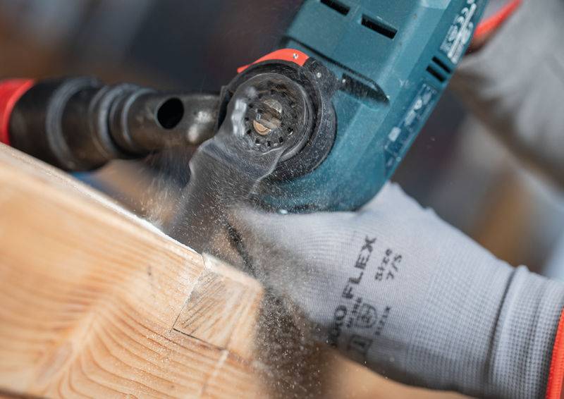 Eine Person in Handschuhen benutzt ein Elektrowerkzeug, um ein Stück Holz zu schneiden und erzeugt dabei Sägespäne.