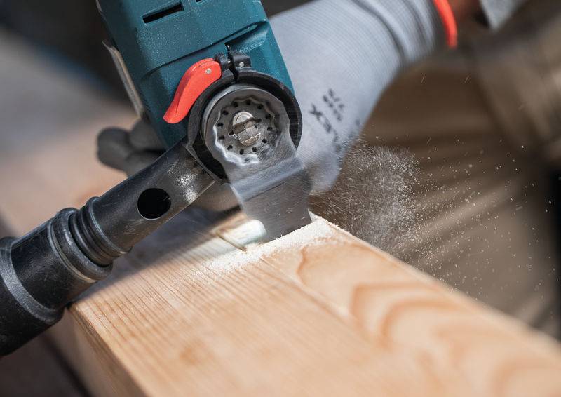 Eine behandschuhte Hand bedient einen Elektroschleiffer auf einer Holzoberfläche und glättet das Holz. Staubpartikel sind in der Luft sichtbar und deuten auf aktives Schleifen hin.