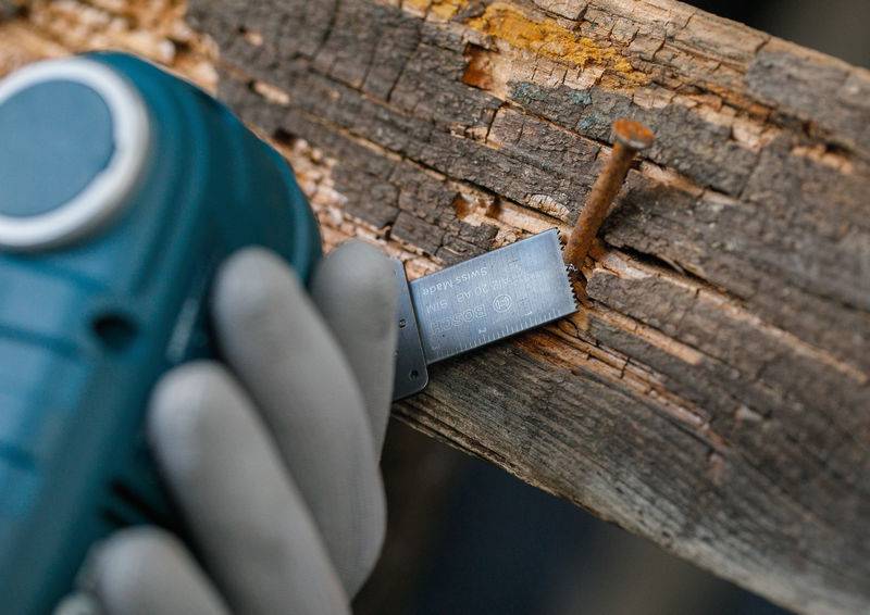 Eine behandschuhte Hand hält ein blaues tragbares Elektrowerkzeug, das ein Holzbrett schneidet, wobei der Fokus auf einem rostigem Nagel liegt, der im Holz steckt.