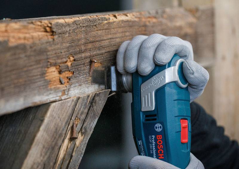 Eine Person mit Handschuhen benutzt ein Bosch-Elektrowerkzeug, um in einen verwitterten Holzbalken zu schneiden, was auf eine Bau- oder Renovierungsaufgabe hinweist.