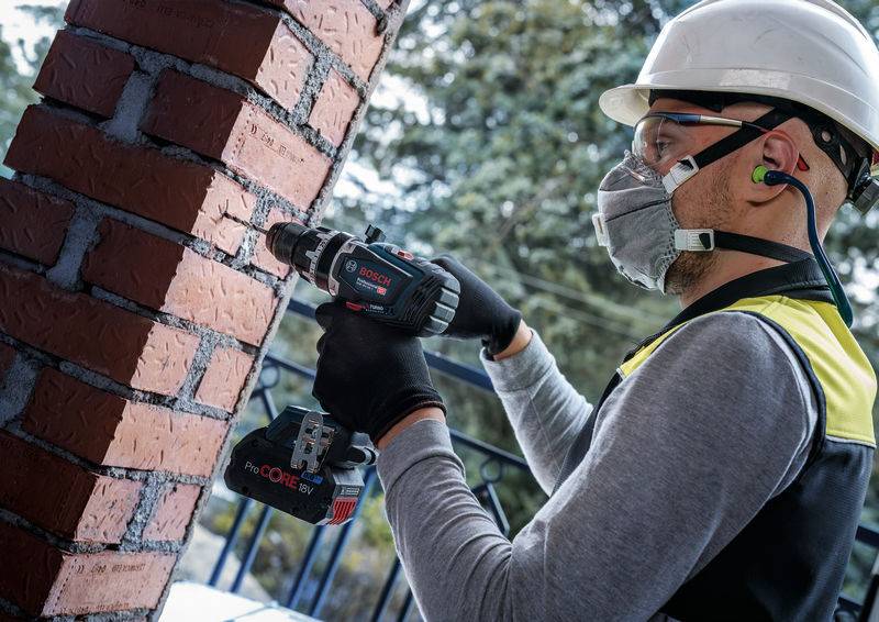 Ein Bauarbeiter in Schutzausrüstung benutzt eine Elektrobohrmaschine an einer Backsteinmauer im Freien und konzentriert sich intensiv auf die Aufgabe.