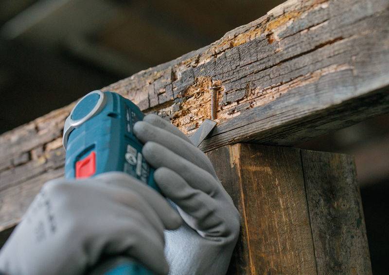 Eine Person mit Handschuhen verwendet ein blaues Elektrowerkzeug zum Schleifen eines abgenutzten, splitternden Holzstücks und glätet dabei die Oberfläche.