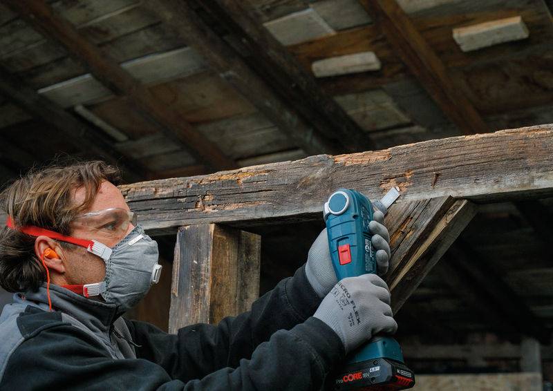 Eine Person in Schutzausrüstung verwendet ein blau-rotes Elektrowerkzeug, um einen Holzbalken in einer rustikalen, unfertigen Struktur zu schleifen.