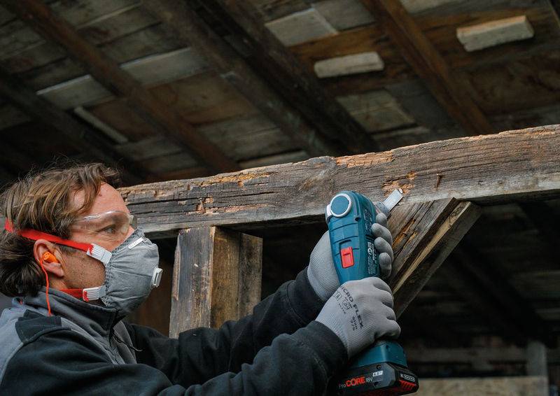 Eine Person in Schutzausrüstung verwendet ein Elektrowerkzeug zum Schleifen eines Holzbalkens in einer rustikalen Umgebung.