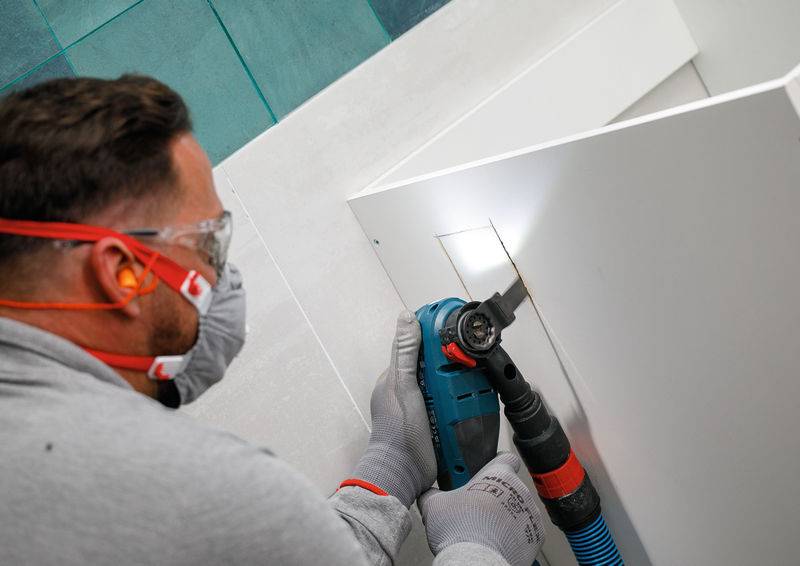 Eine Person in Schutzausrüstung nutzt ein Elektrowerkzeug, um ein quadratisches Loch in ein weißes Panel zu schneiden, wobei eine Leuchte den Arbeitsbereich beleuchtet.