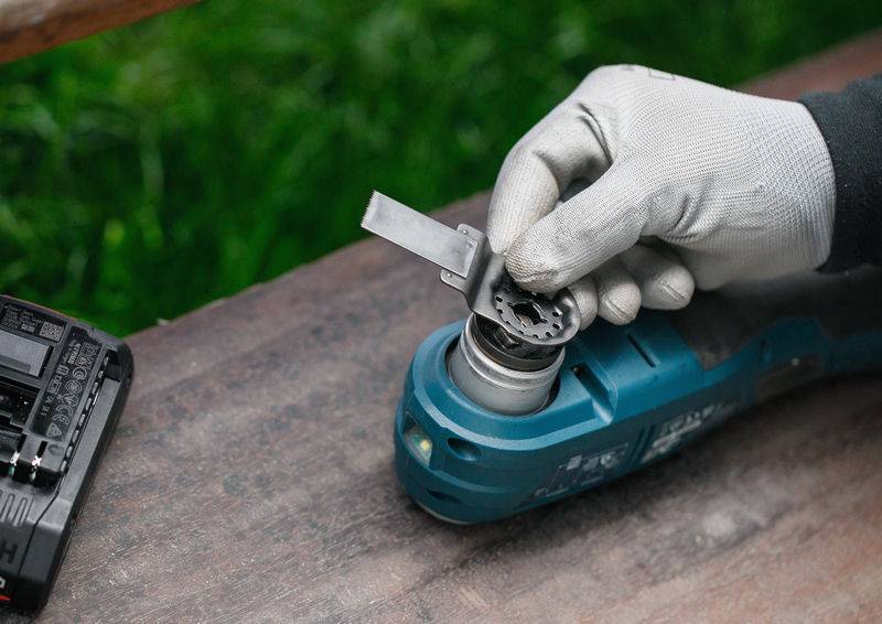 Eine Person mit Handschuhen wechselt ein Blatt an einem oszillierenden Multiwerkzeug, das auf einer Holzoberfläche im Freien liegt.