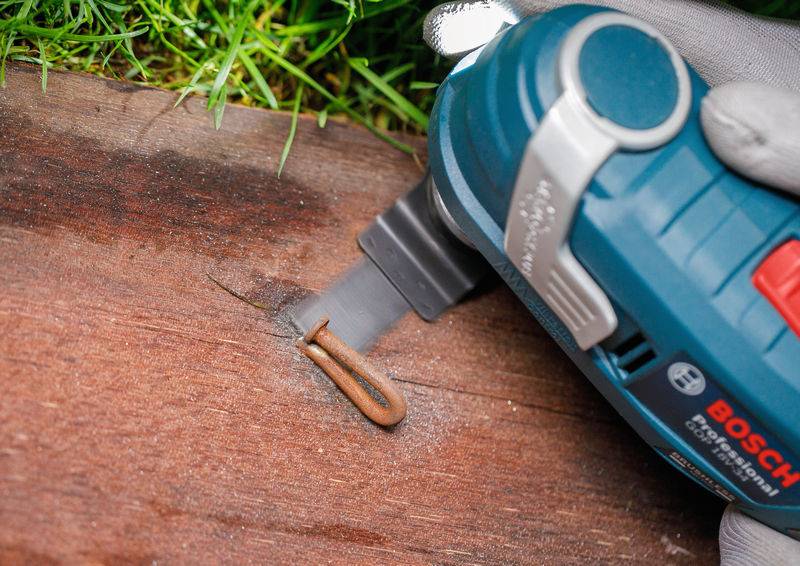 Ein Bosch-Elektrowerkzeug wird verwendet, um einen in einem Holzbrett eingebetteten Metallheftklammerung durchzusägen. Gras ist im Hintergrund sichtbar.