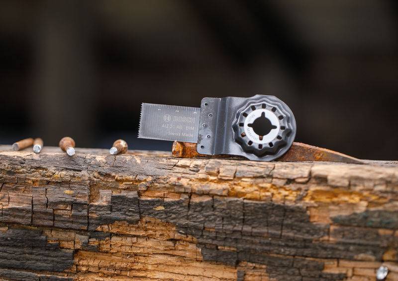 Eine Multitool-Klinge und mehrere Nägel liegen auf einer rauen, verwitterten Holzoberfläche, was auf einen Werkzeuggebrauch-Kontext hindeutet.