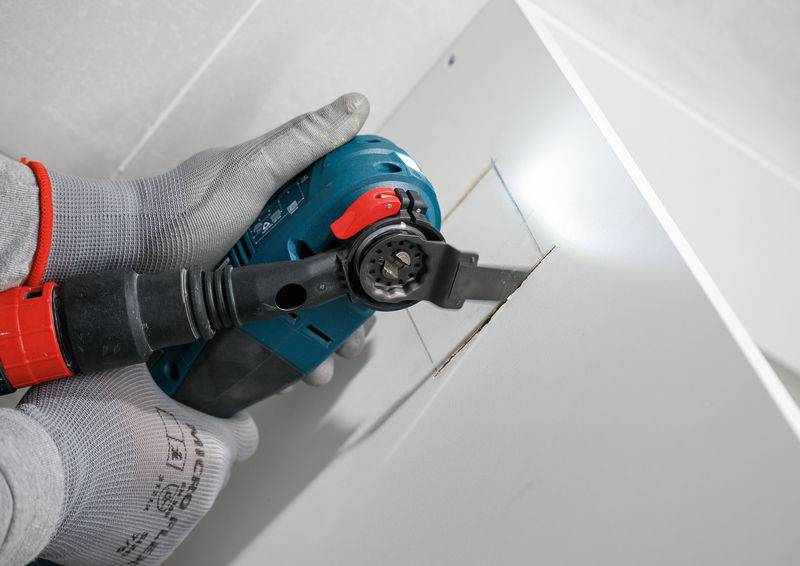 Eine Person mit Handschuhen verwendet ein Elektrowerkzeug, um ein rechteckiges Loch in ein weißes Panel zu schneiden, wobei sie sich auf Präzision und Kontrolle konzentriert.