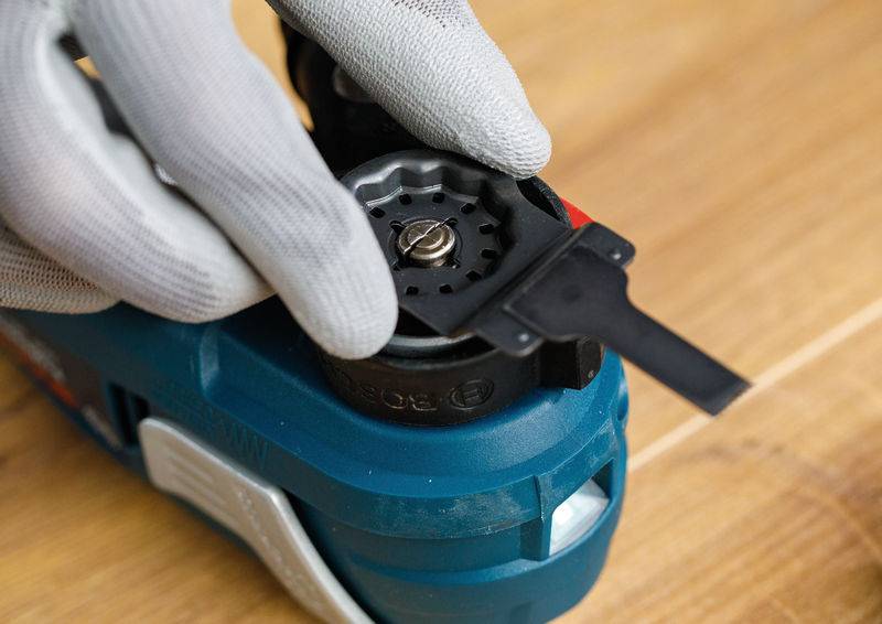 Eine Person, die Handschuhe trägt, benutzt ein elektrisches Elektrowerkzeug auf einer Holzoberfläche und führt eine Zubehörklinge in den Schlitz des Werkzeugs ein.