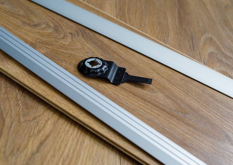 Ein Metallwerkzeug ruht auf einem holzähnlichen Laminatboden neben einer Messlatte, was auf Bau- oder Installationsarbeiten hindeutet.