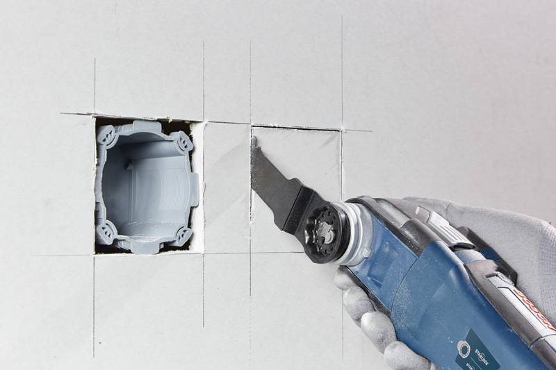 Eine Person verwendet ein oszillierendes Multifunktionswerkzeug, um ein quadratisches Loch in Trockenbau für eine Elektroinstallationsdose zu schneiden, wobei ein Heimwerkerprozess demonstriert wird.