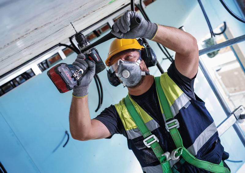Arbeiter in Bauausrüstung, einschließlich Helm, Maske und Sicherheitsgurt, verwendet einen Elektroschrauber an einer Decke in einem Gebäudeinneren.