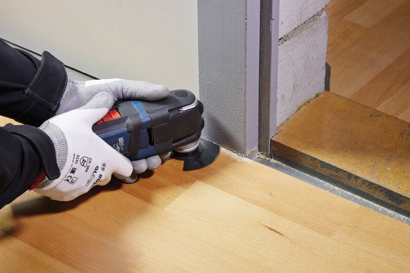 Eine Person nutzt ein elektrisches Multifunktionswerkzeug mit Oszillation, um eine Holzfusskante neben einer Türöffnung zu trimmen, während sie Schutzhandschuhe trägt.