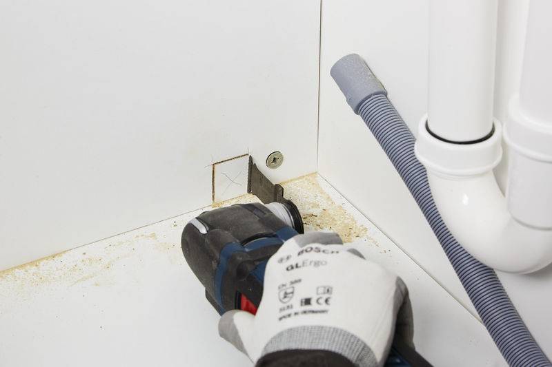 Ein Arbeiter, der ein Elektrowerkzeug benutzt, schneidet ein quadratisches Loch in weiße Möbel in der Nähe von Rohren, möglicherweise für die Installation oder Wartung von Sanitäranlagen.