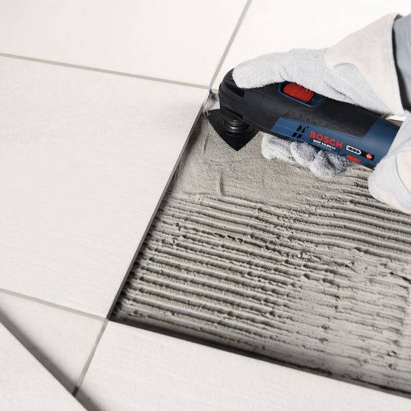 Eine Person verwendet ein Bosch-Oszillationswerkzeug, um Fugenmaterial zwischen weißen Bodenfliesen zu entfernen und zeigt damit Fliesen-Reparatur- oder Wartungsarbeiten.