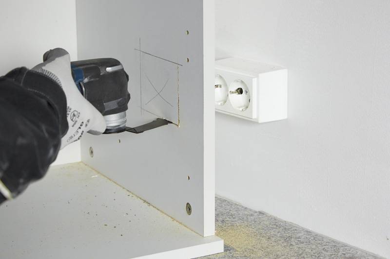 Eine Person, die mit einem Elektrowerkzeug ein quadratisches Loch in eine weiße Holzplatte schneidet, mit einer Steckdose in der Nähe an der Wand.