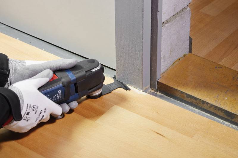 Eine Person mit Handschuhen nutzt eine Multifunktions-Säge, um ein Holzbodenplatte an der Kante nahe einer Wand zu schneiden und demonstriert dabei präzises Schneiden.