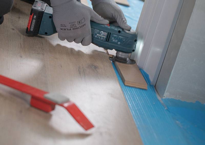 Eine Person in Handschuhen verwendet ein Elektrowerkzeug, um Bodenbelag in der Nähe einer Wand zu schneiden. Ein Holzbrett und Bodenbelag-Installationswerkzeuge sind sichtbar.