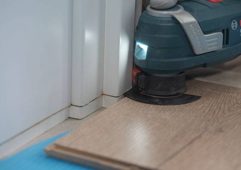 Ein Elektrowerkzeug mit einer kreisförmigen Schneideklinge trimmt ein Holzparkett neben einer weißen Wand, während ein Licht den Bereich beleuchtet.